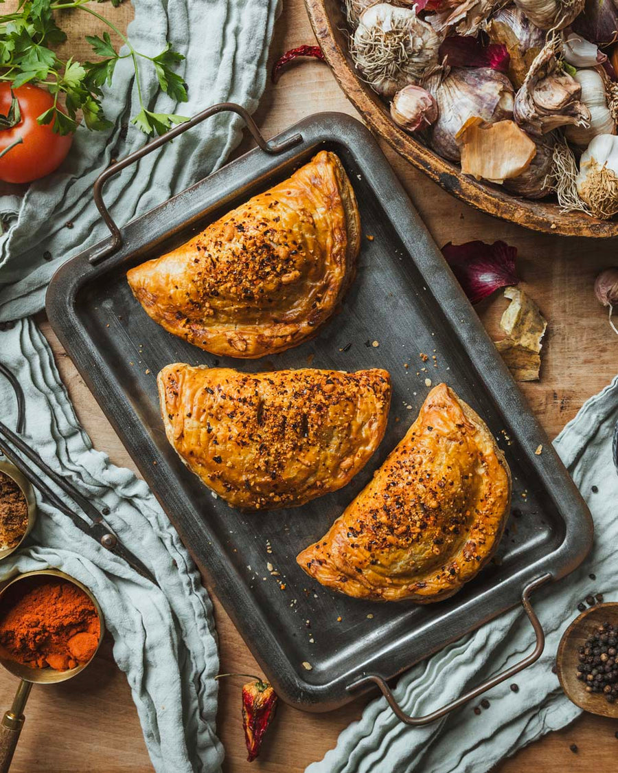 Three BBQ pulled pork pasties on a baking sheet with ingredients around them, including tomatoes, garlic, and spices.