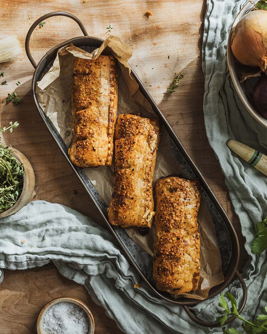 Three BBQ pulled pork sausage rolls on a metal tray with a wooden background, garnished with herbs and spices.
