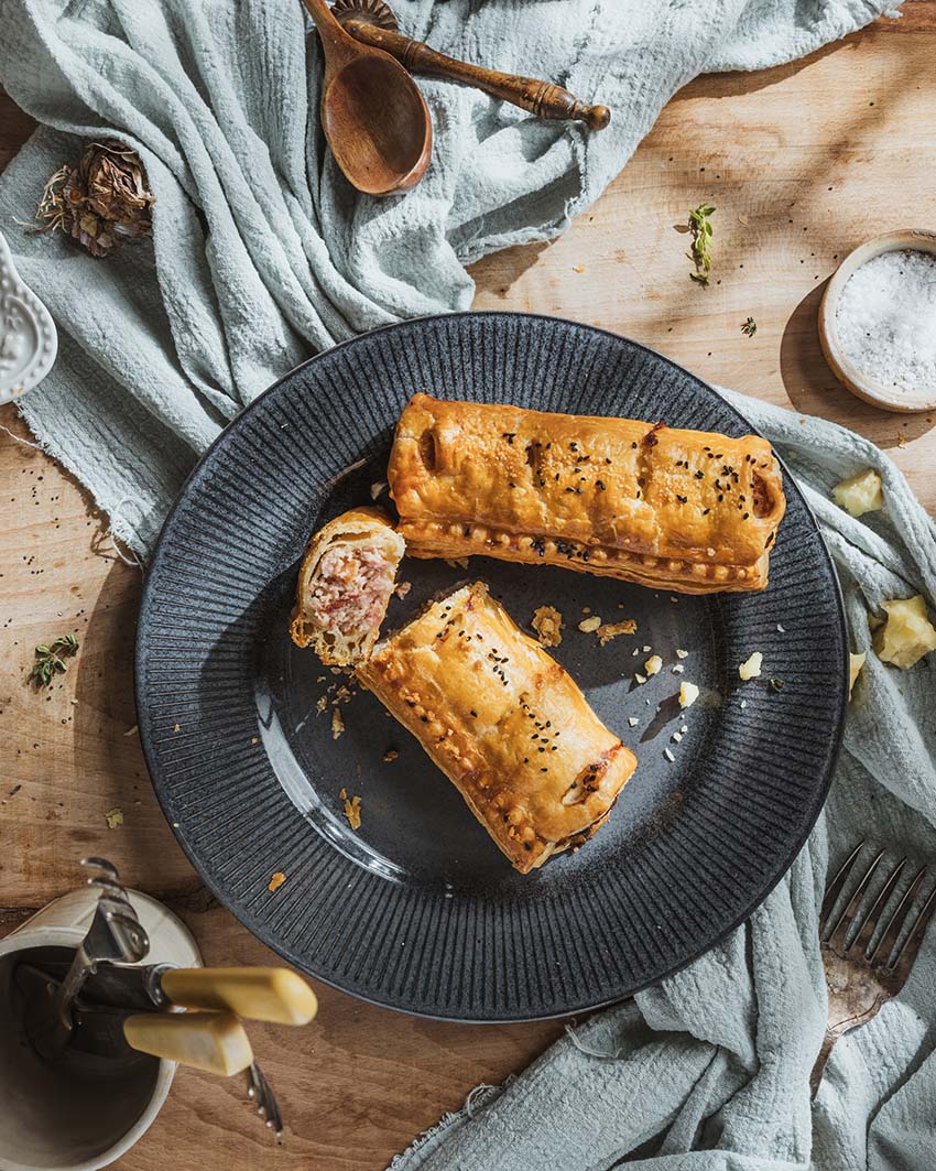Two bacon and mature cheddar sausage rolls on a black plate with one cut to show the filling, served on a wooden table with a blue cloth and decorative items in the background.
