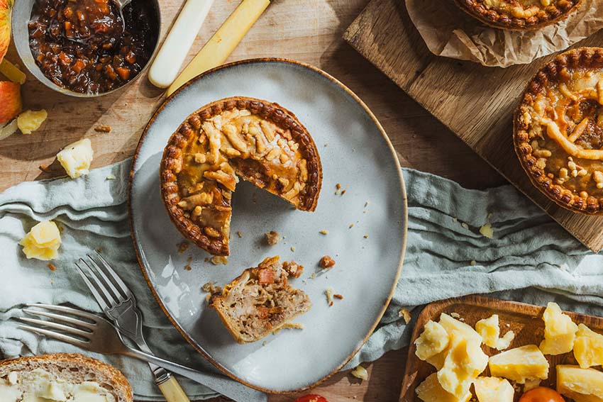 One Cheese & Pickle Pork Pie cut open showing pork, cheese and pickle filling. Two other Pork pies on a wooden chopping board 