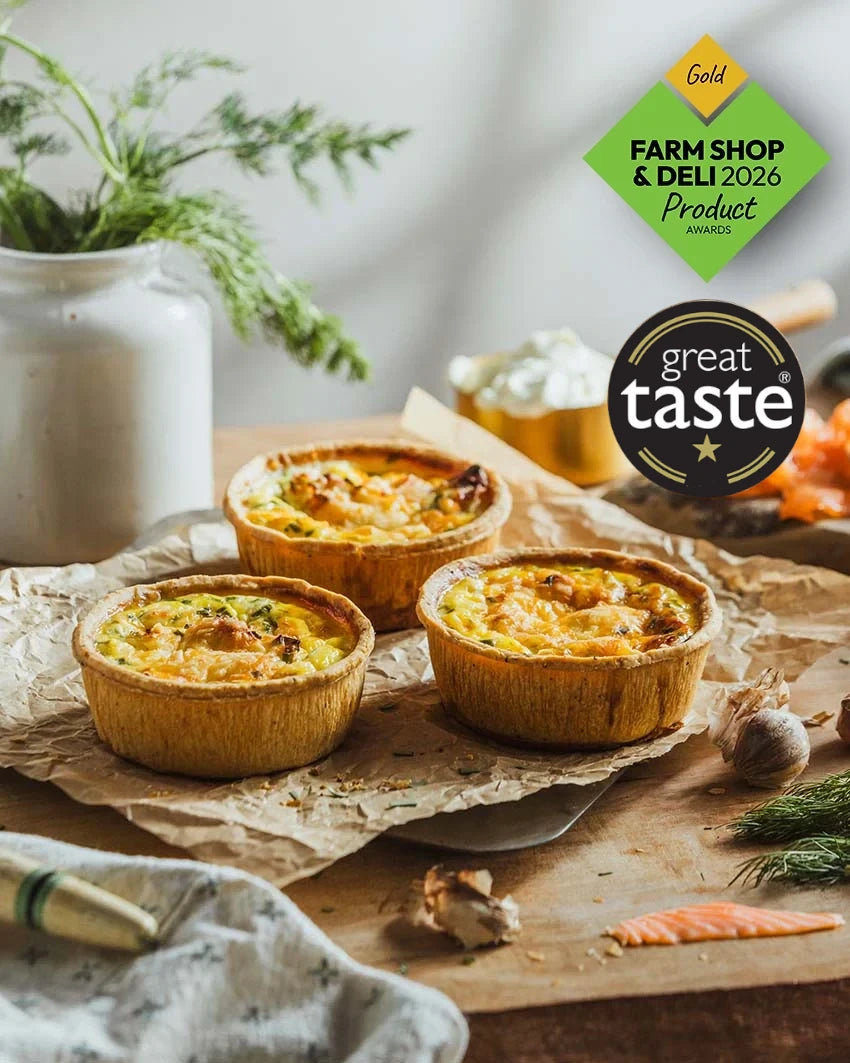 Three baked Smoked Salmon quiches in  arranged on parchment paper on a wooden board, with a white ceramic jar, herbs, garlic, and salmon garnish around them. Two promotional badges appear on the right: a green Farm Shop & Deli 2026 Product Awards gold badge and a black Great Taste award badge.